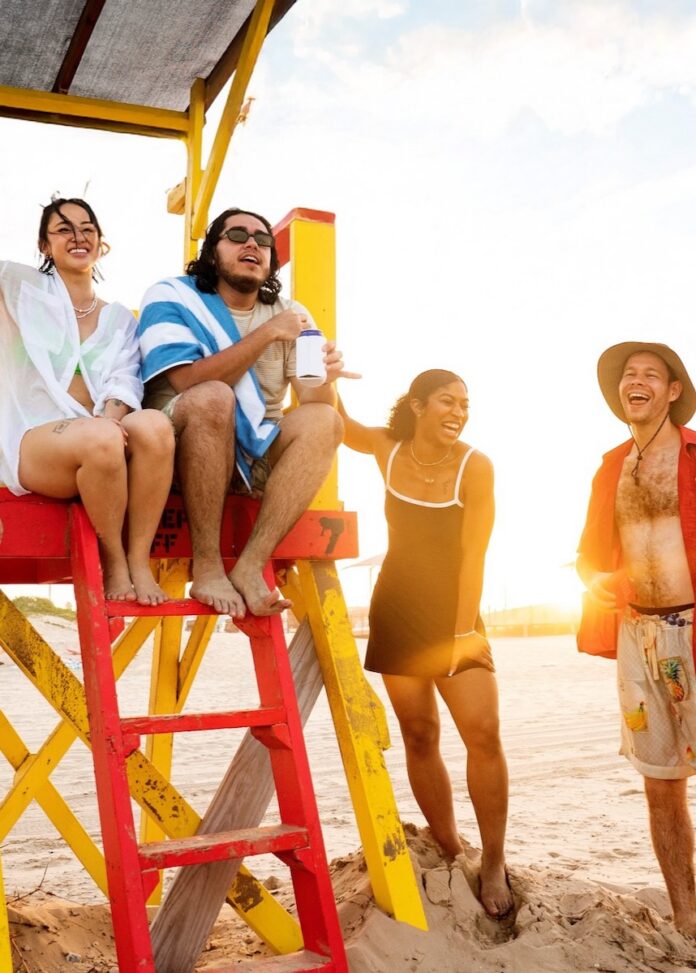 A couple sits on a lifeguard stand as another couple stands beside it on the beach.