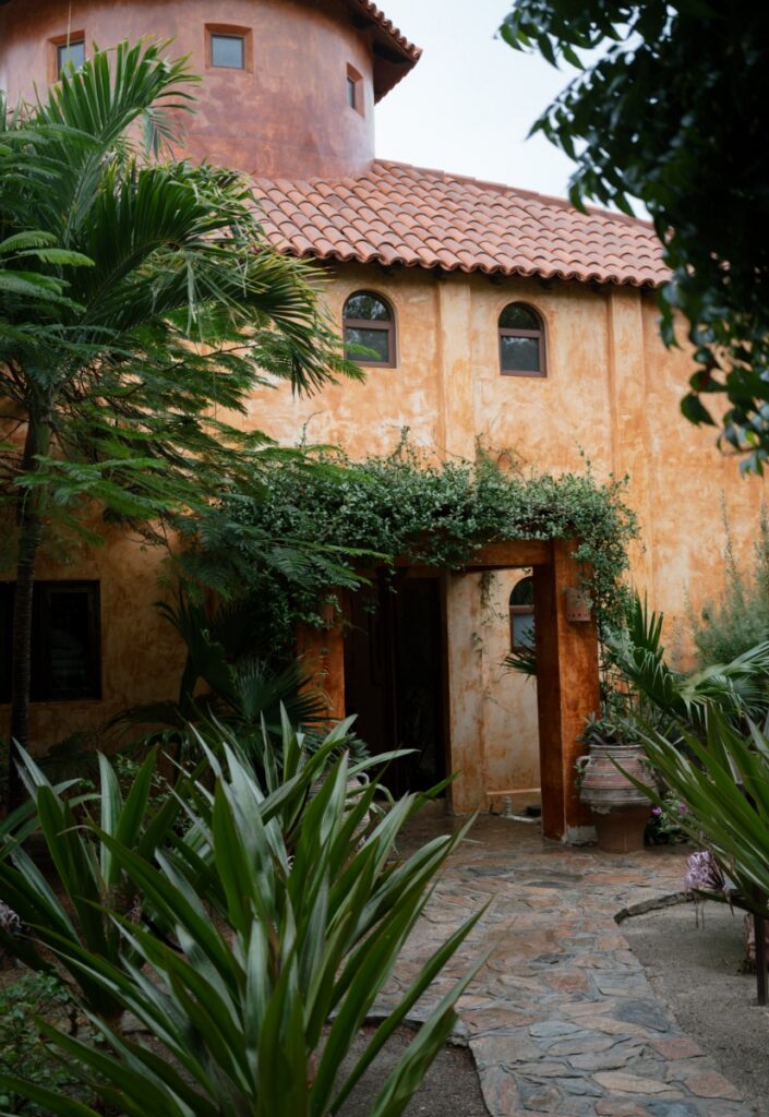 The exterior of Villa Santa Cruz in Todos Santos, Mexico, featuring traditional terracotta roof tiles and lush tropical greenery along a stone path.