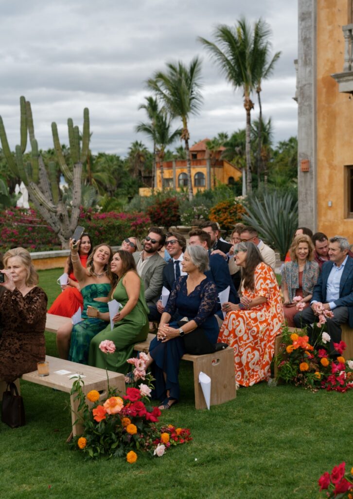 Wedding guests seated on wooden benches on the lush lawn of Villa Santa Cruz during an outdoor ceremony in Todos Santos, Mexico.