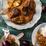 A white plate of Andhra-Style Stuffed Eggplant Curry with large eggplants throughout, and a dish of rice in the bottom right corner.