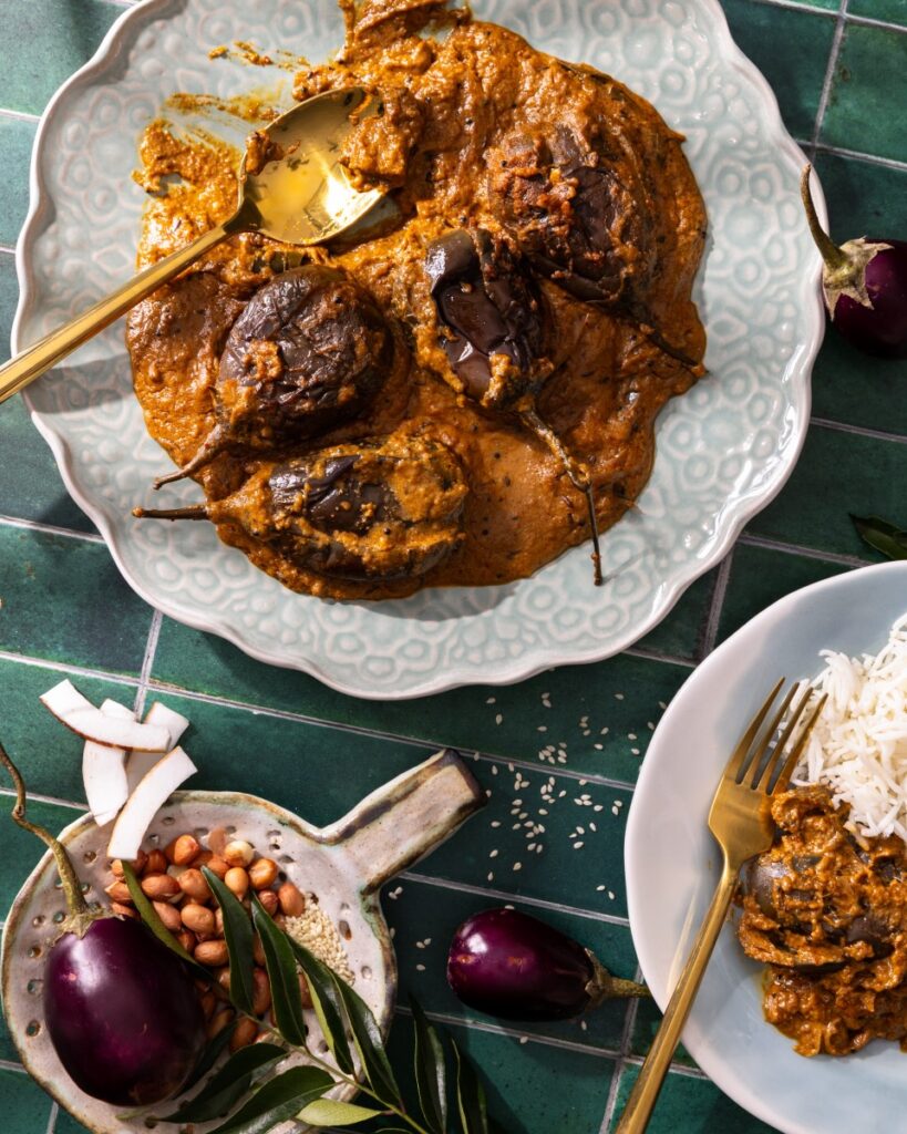 A white plate of Andhra-Style Stuffed Eggplant Curry with large eggplants throughout, and a dish of rice in the bottom right corner.