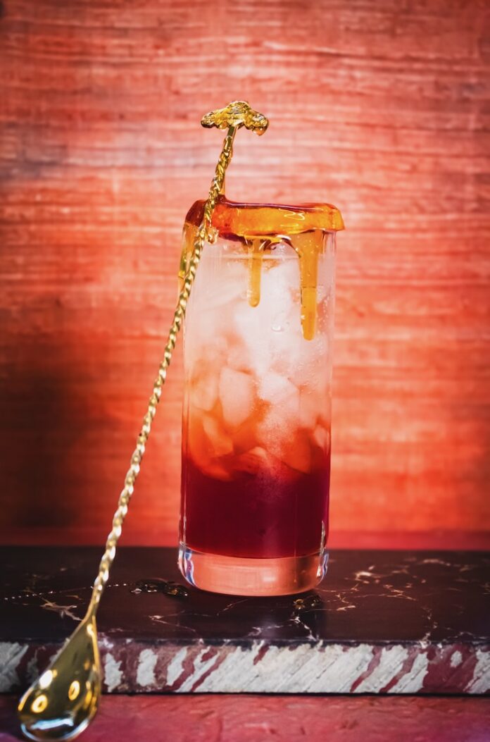 A tall cocktail in red with a honey blood orange garnish over top of the glass and honey running down the sides of the glass as a golden bar spoon sits against it.