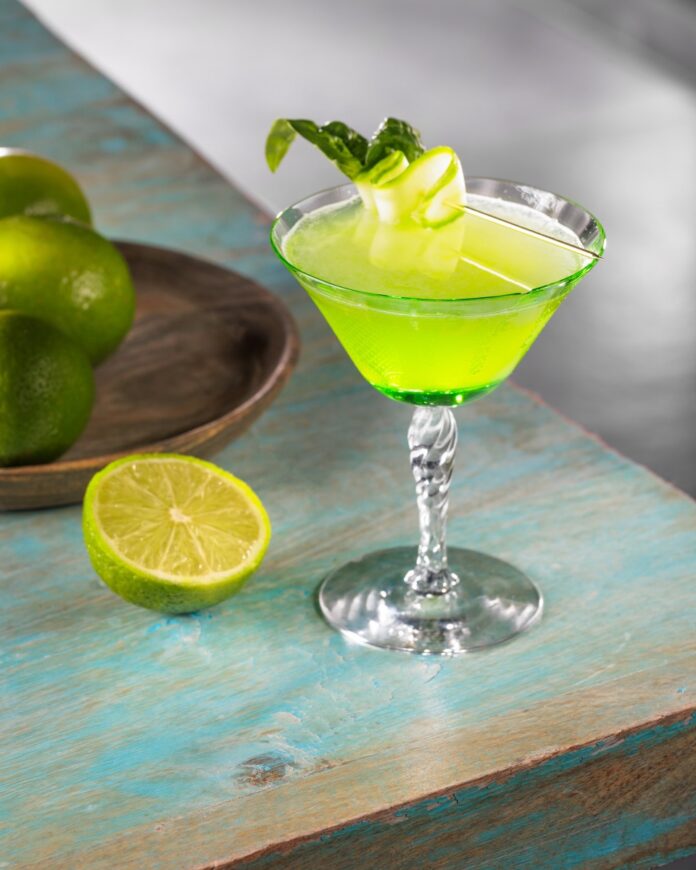 A green basil and cucumber gimlet in a Nick and Nora glass with half a lime sitting beside it.
