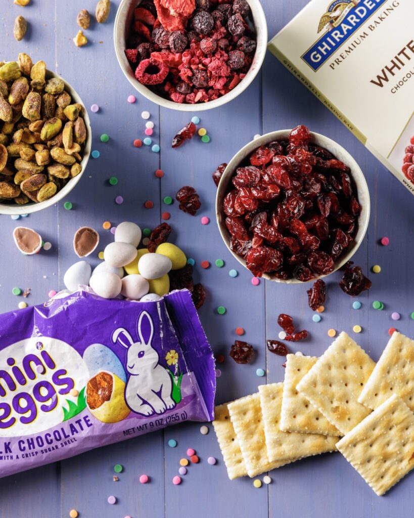 A purple table with small bowls of ingredients like saltine crackers, mini eggs, dried berries, and pistachios.