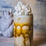 A white chocolate caramel frozen mudslide in a tall glass with whipped cream on top and a white anatomical heart sculpture behind it.