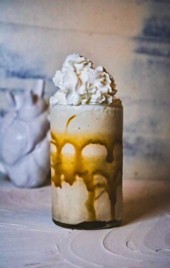 A white chocolate caramel frozen mudslide in a tall glass with whipped cream on top and a white anatomical heart sculpture behind it.