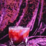 A blackberry bramble sits in a rocks glass on top and in front of purple velvet.