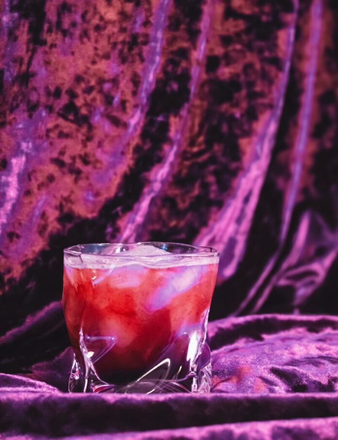 A blackberry bramble sits in a rocks glass on top and in front of purple velvet.