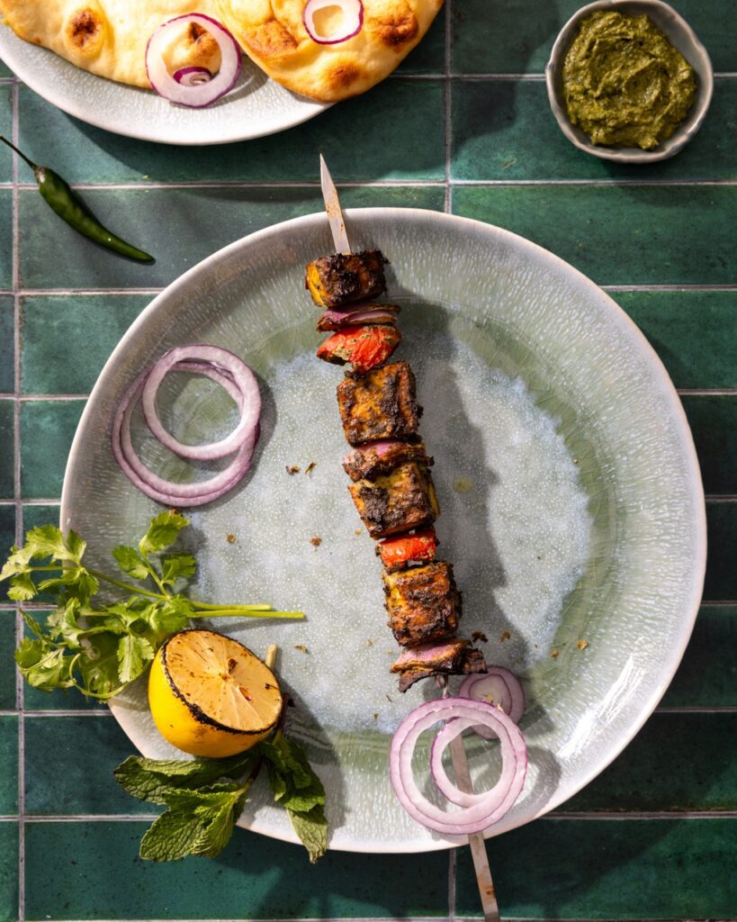 A plate with a singular Hariyali Paneer Kebab on it, rings of onions, and a grilled lemon.