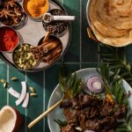A plate of Kerala-Style Dry Beef Stir-Fry on a green plate surrounded by herbs while a circular tray of spices sits adjacent to it.