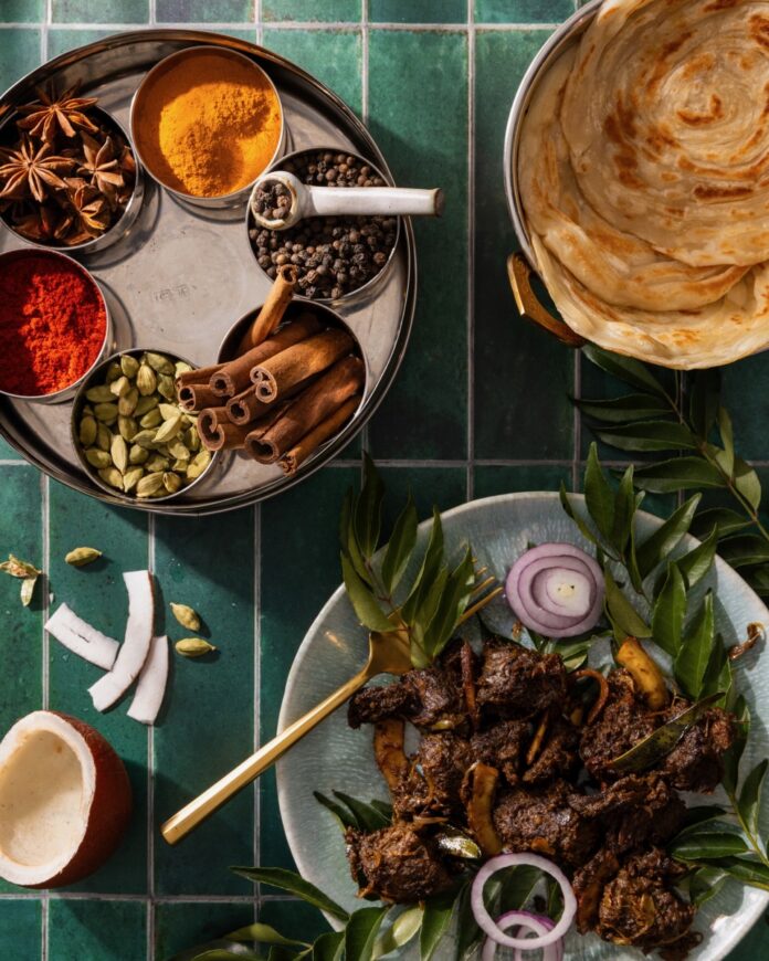 A plate of Kerala-Style Dry Beef Stir-Fry on a green plate surrounded by herbs while a circular tray of spices sits adjacent to it.