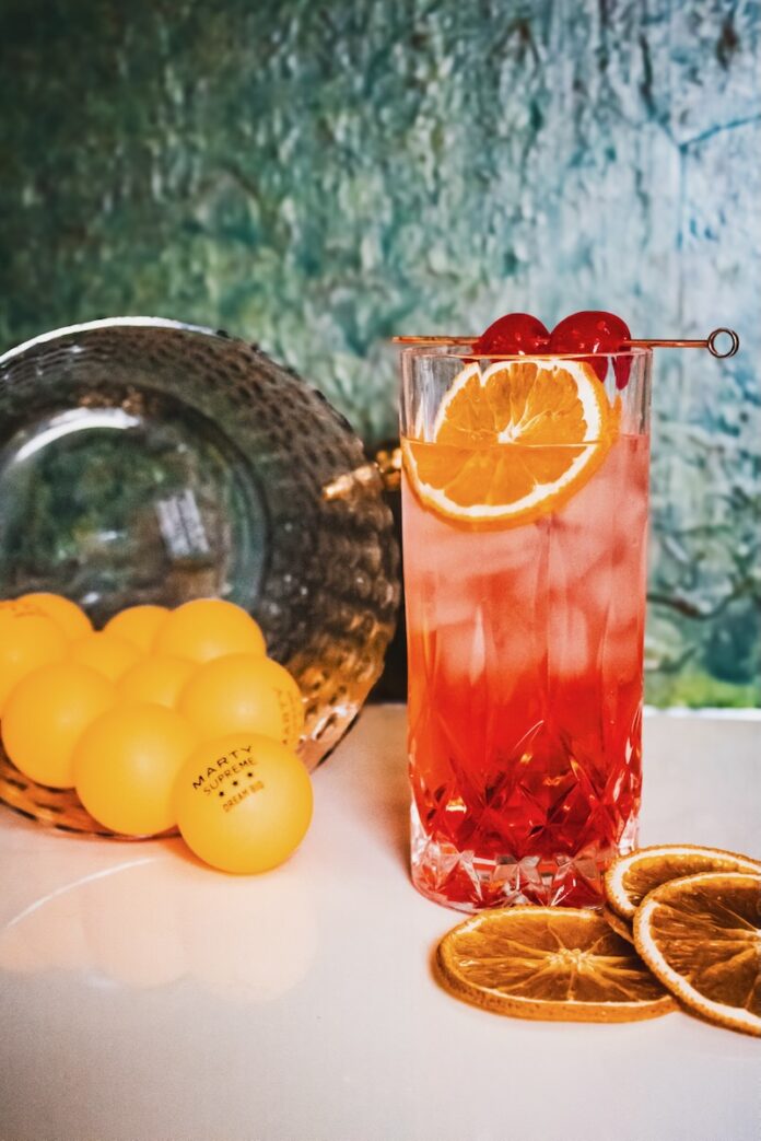 An Orange Dirty Shirley Cocktail sits beside a spilled bucket of ping-pong balls with cherries and an orange wheel for garnish.