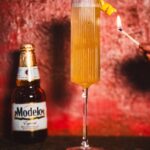 A person holds a match to a flute French 75 with a lemon twist and a bottle of Modelo in the background.