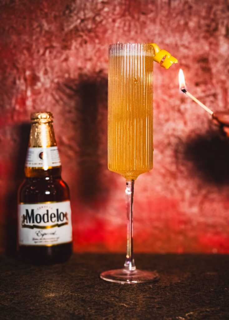 A person holds a match to a flute French 75 with a lemon twist and a bottle of Modelo in the background.