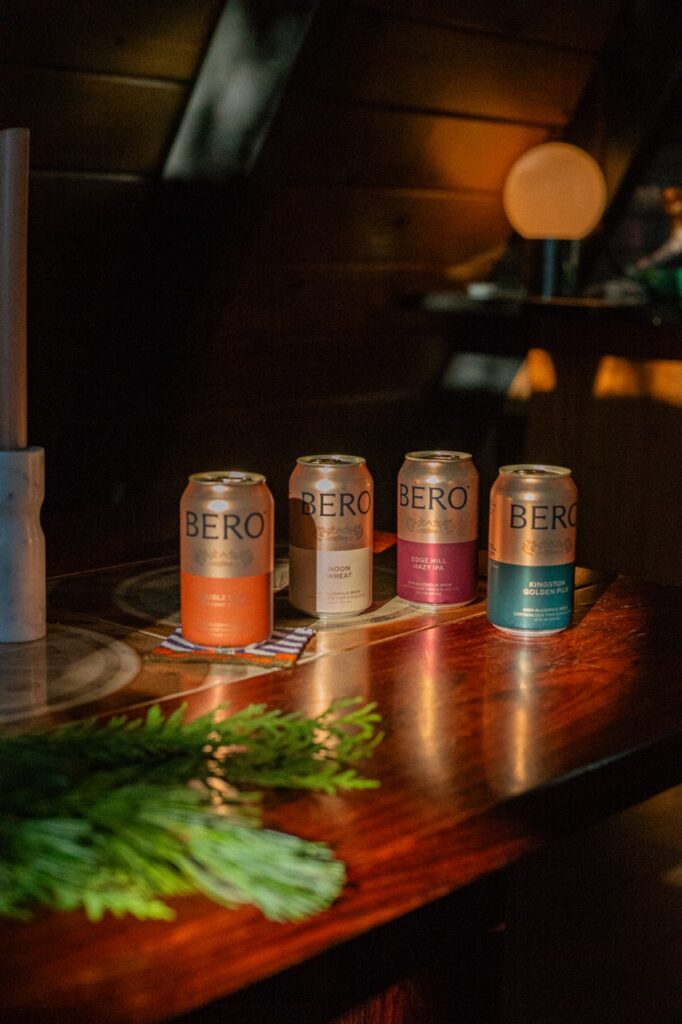 Four cans of BERO non-alcoholic beer sit on a wooden table with a green palm in front of it.