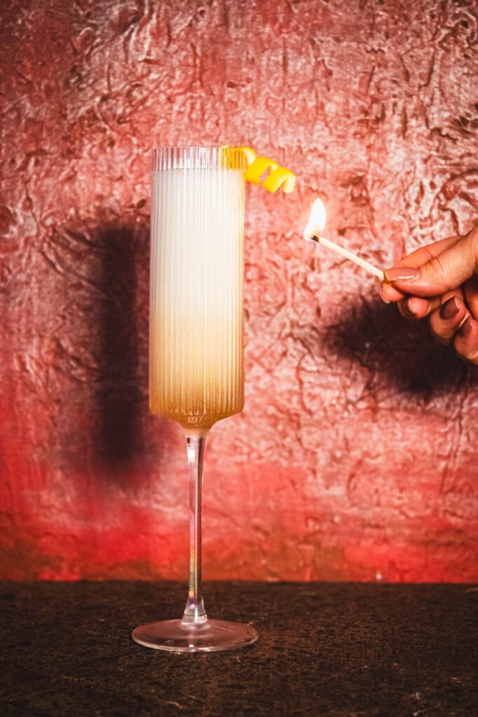 A person holds a match to a flute cocktail with a lemon twist garnish.