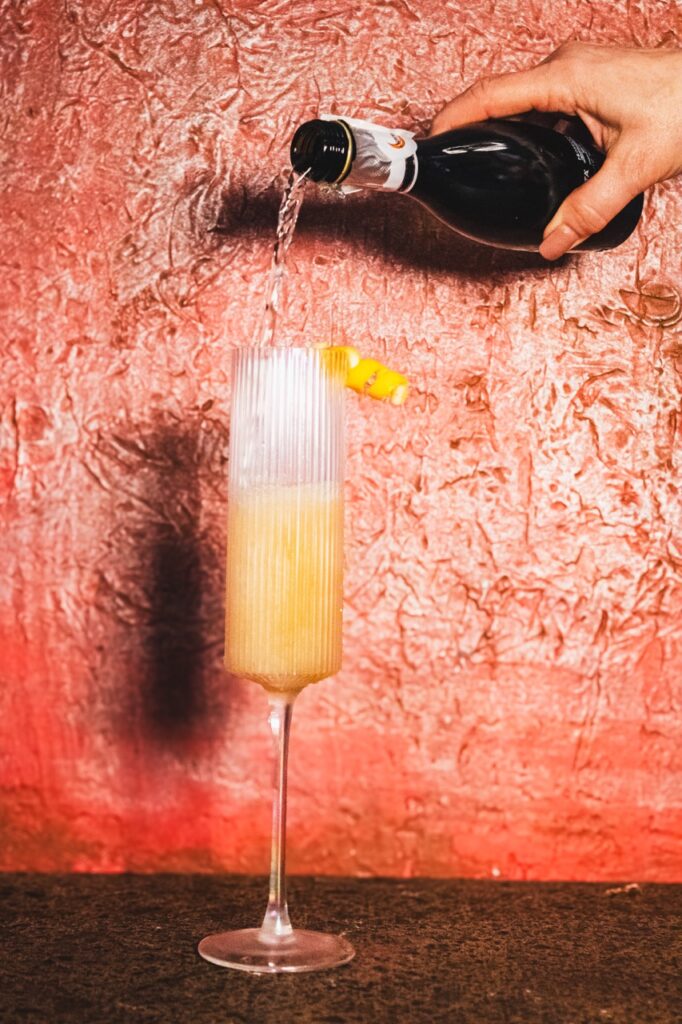 A person pours champagne into a cocktail flute with a lemon twist garnish.