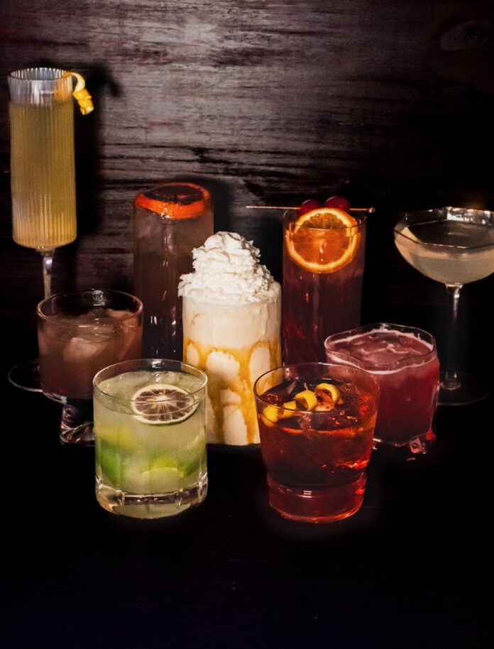 A dark table full of cocktails in short and tall glasses arranged with fruit garnishes and a mudslide in the middle.