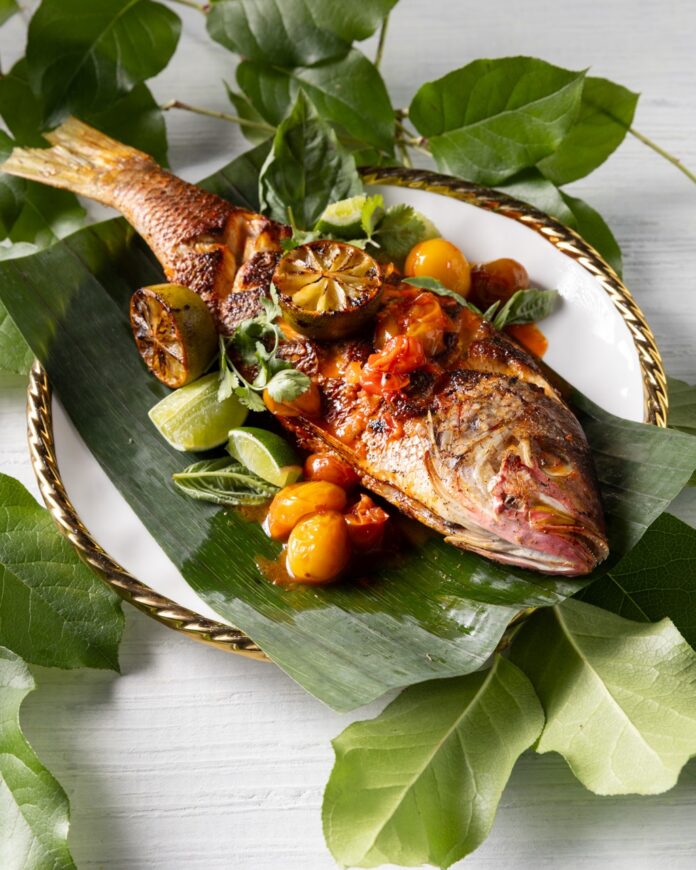 A white plate with leaves every where and a red snapper laying on the plate with taro root smash and salsas.