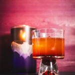 A whiskey and beet cocktail for Sinners sits on a table in front of a bat candle and barn door.