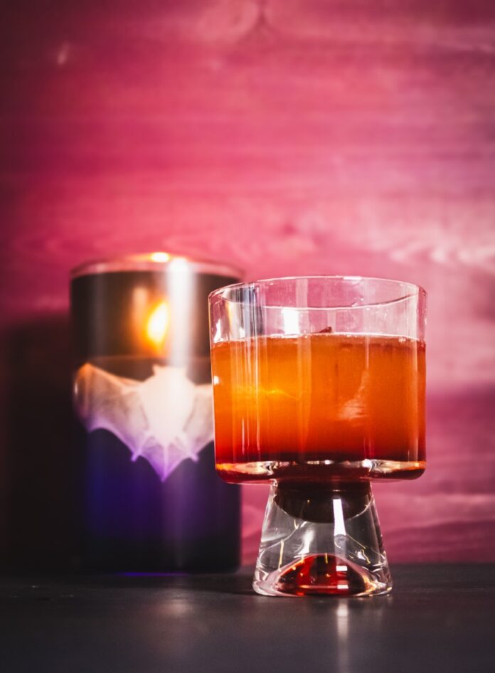 A whiskey and beet cocktail for Sinners sits on a table in front of a bat candle and barn door.