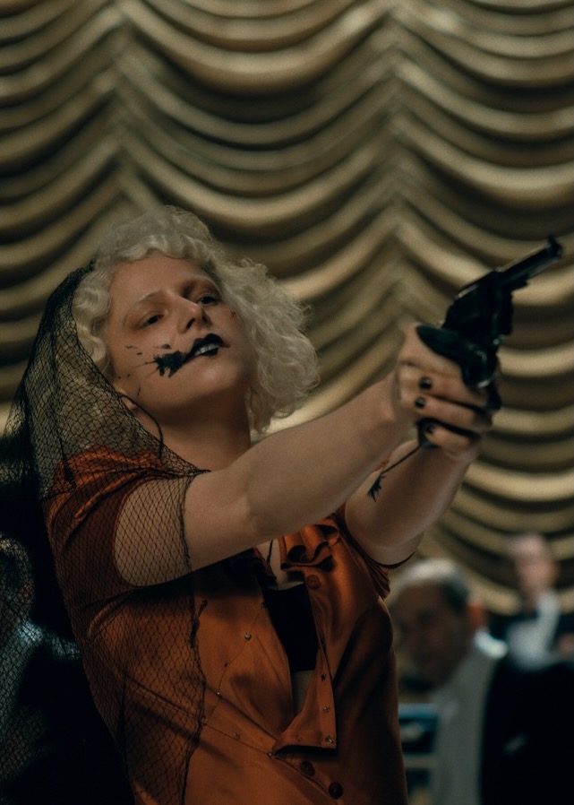 The Bride holds a gun against curtains of gold.