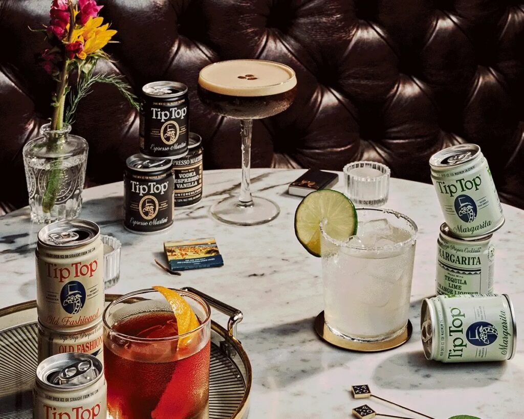 Canned cocktails on a table sit beside glasses of cocktails like an Espresso Martini.