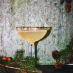 A cocktail in a coupe glass sits on a green background with sprigs of pine trees peeking in.