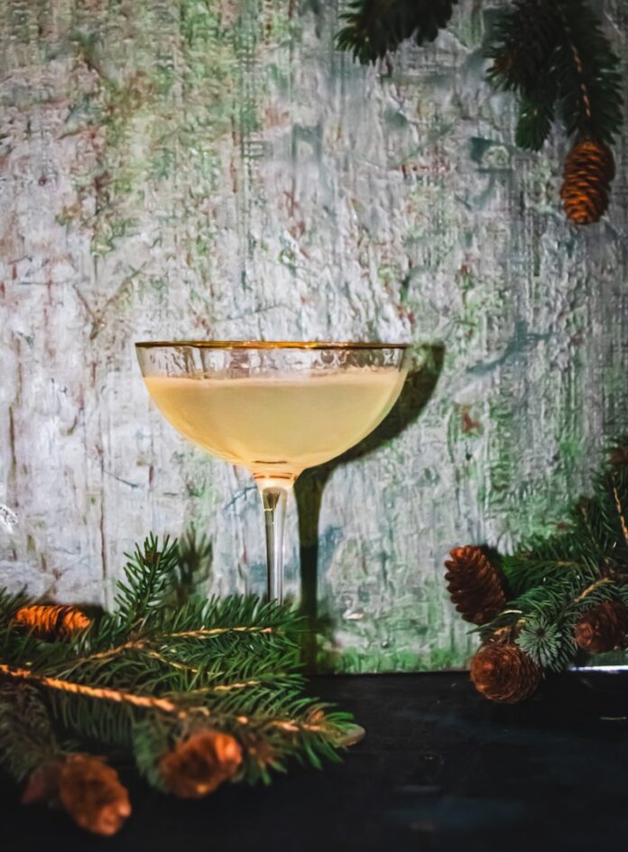 A cocktail in a coupe glass sits on a green background with sprigs of pine trees peeking in.