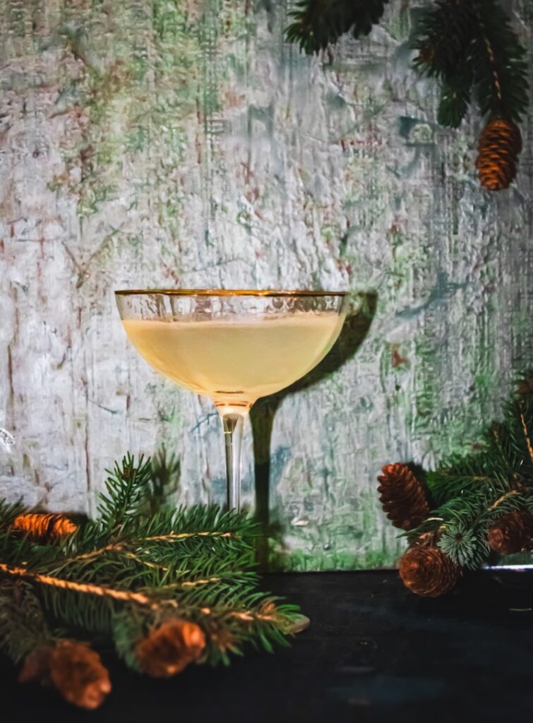 A cocktail in a coupe glass sits on a green background with sprigs of pine trees peeking in.