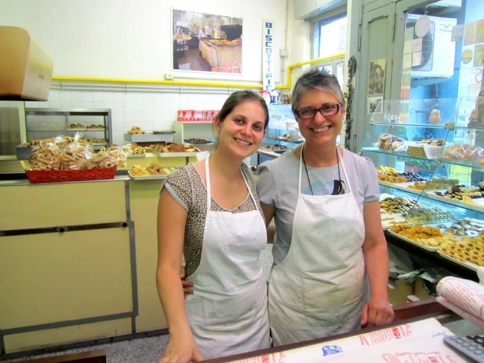 Two women in aprons stand behind a counter.