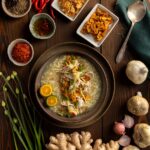A bowl of Arroz Caldo Chicken and Rive Porridge sits on a brown table with small containers of spices all around it.