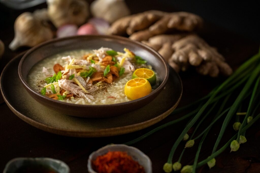 A brown bowl full of Arroz Caldo Chicken and Rice Porridge topped with lemons, greens, and other seasonings.