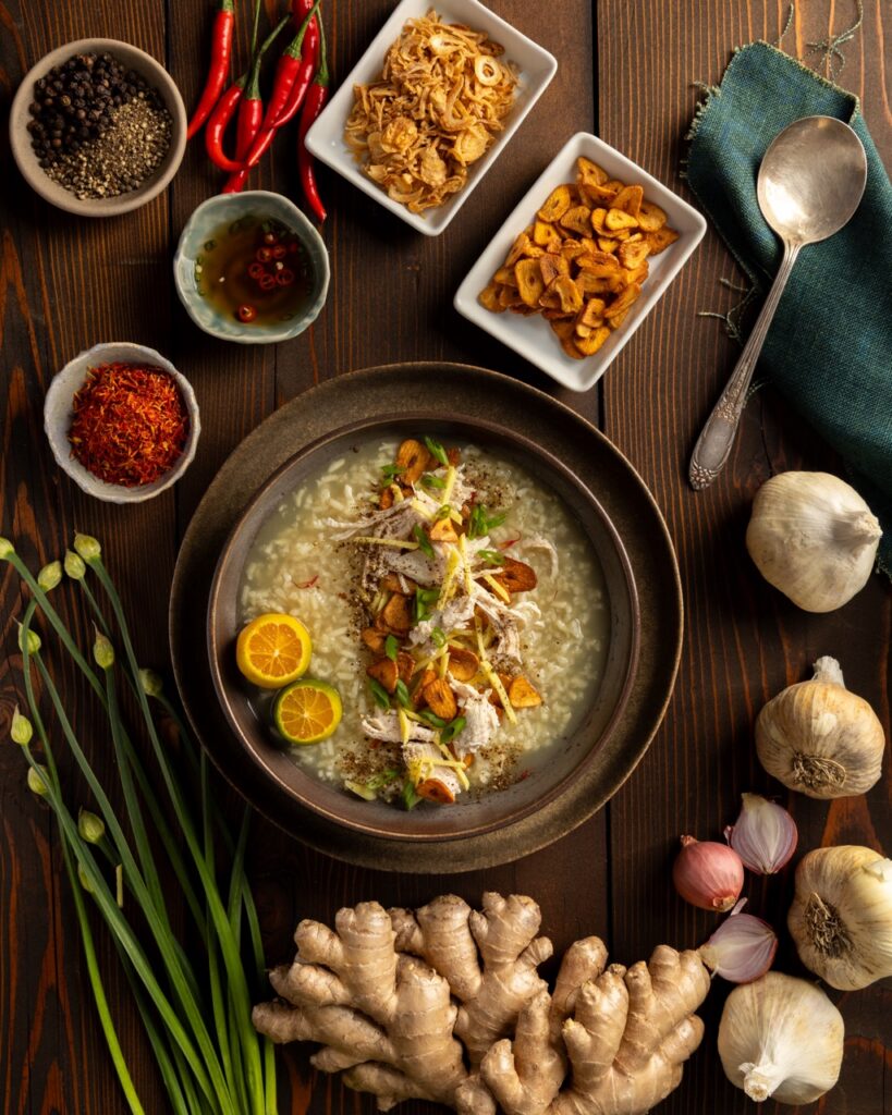 A bowl of Arroz Caldo Chicken and Rive Porridge sits on a brown table with small containers of spices all around it.