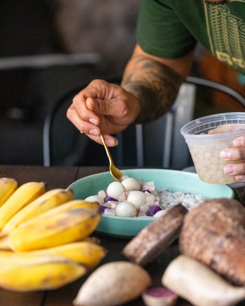 A man spoons ingredients into a pale green bowl full of porridge with bananas and sweet potatoes in front of the bowl.