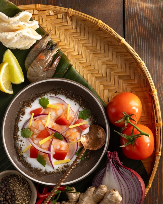 A small bowl of Kinilaw na Hipon (Filipino Ceviche) sits with a spoon in it on a woven tray next to tomatoes and citrus.