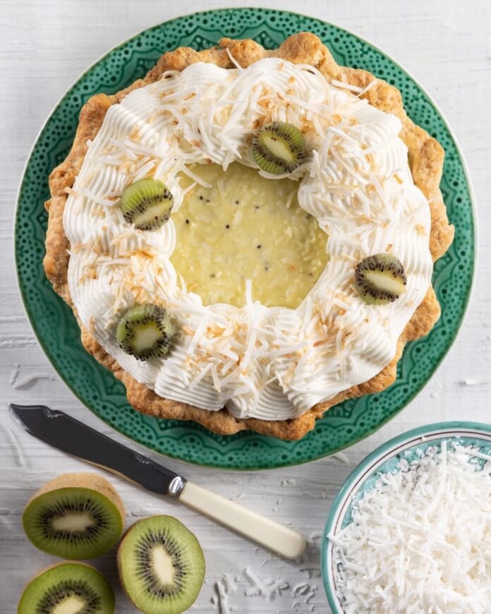 A kiwi-coconut cream pie sits on a green plate with whipped cream, shredded coconut, and kiwi slices on top.