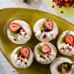 Six round swirls of pavlova with kiwi jam in the center, a piping of orange zest whipped cream, and a half strawberry on top.