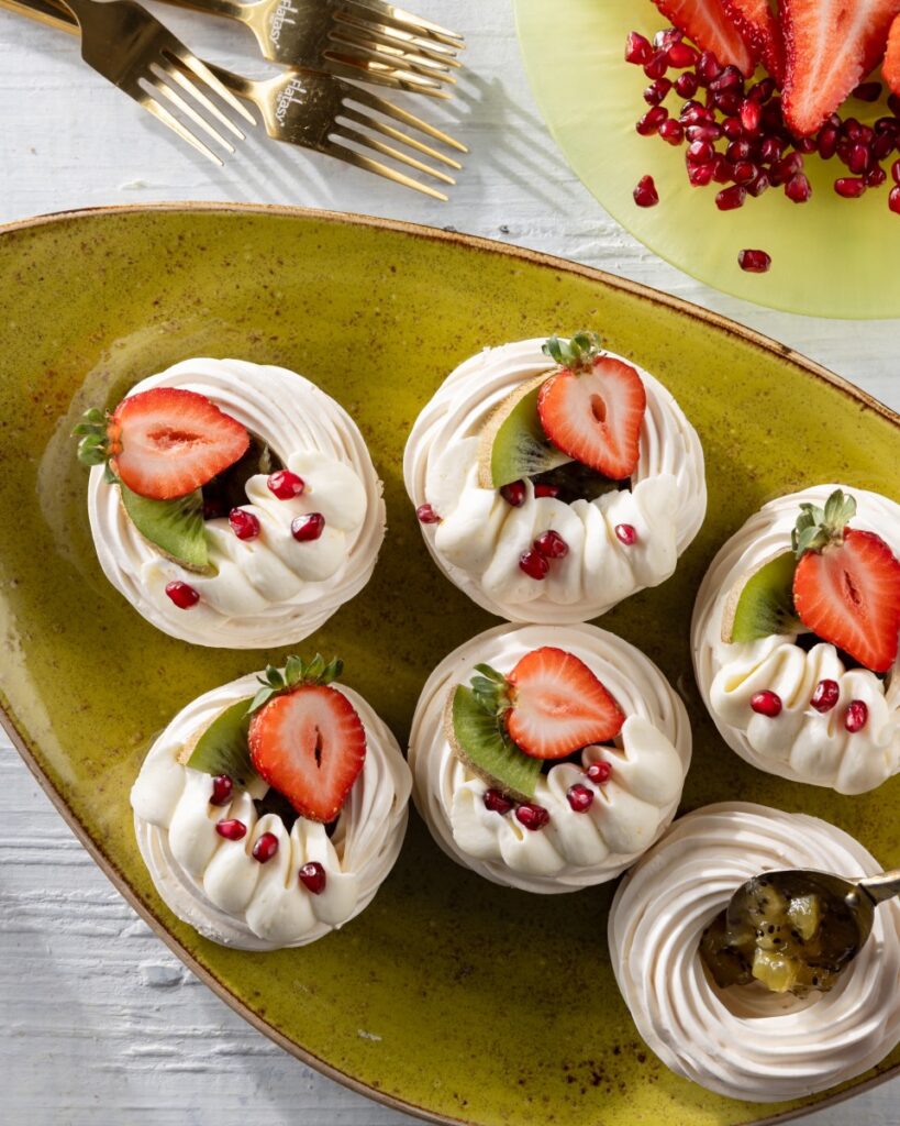 Six round swirls of pavlova with kiwi jam in the center, a piping of orange zest whipped cream, and a half strawberry on top.