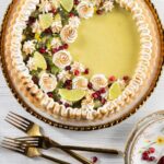 A kiwi and lime tart in a pie tie with meringue, lime wedges, kiwi, and pomegranate seeds on top.