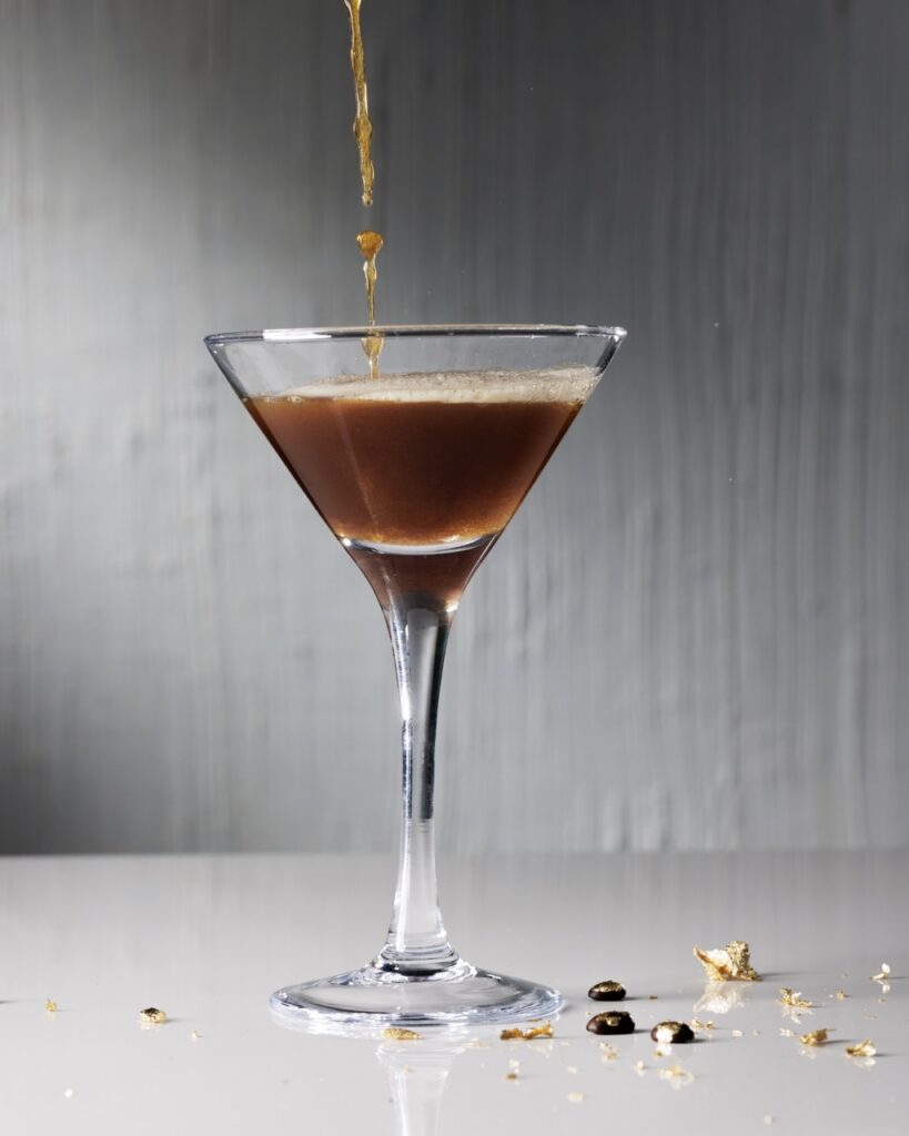Coffee pours into an espresso martini in a tall glass with gold leaf coffee beans below it.