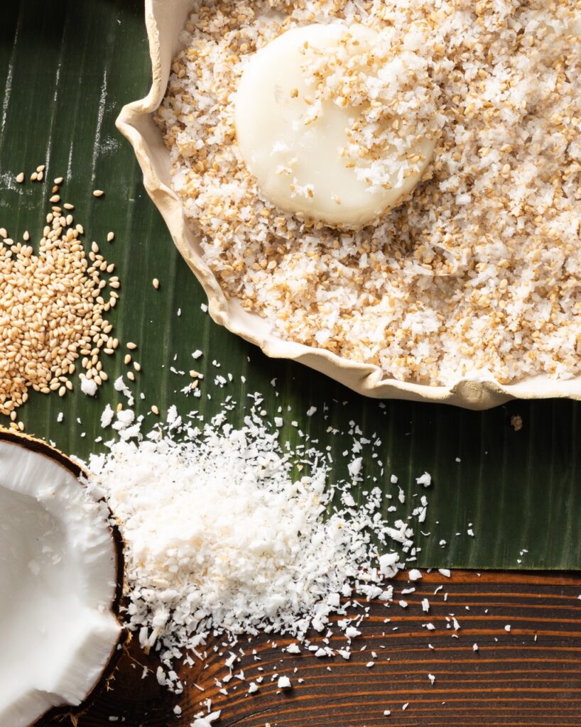 A round ball of chewy rice cake palitaw sits in a bowl fo coconut flakes with toasted sesame seeds and a coconut off to the side.