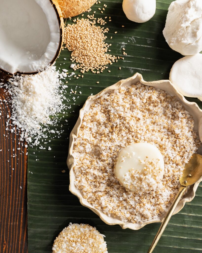 A palitaw chewy rice cake sits in a white bowl of coconut flakes with a coconut and other ingredients in the top left corner.