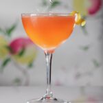 An orange color cocktail in a tall glass with a lemon twist garnish and a floral backdrop behind it.
