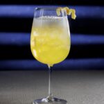 A tall glass with a yellow cocktail with a lemon twist garnish.