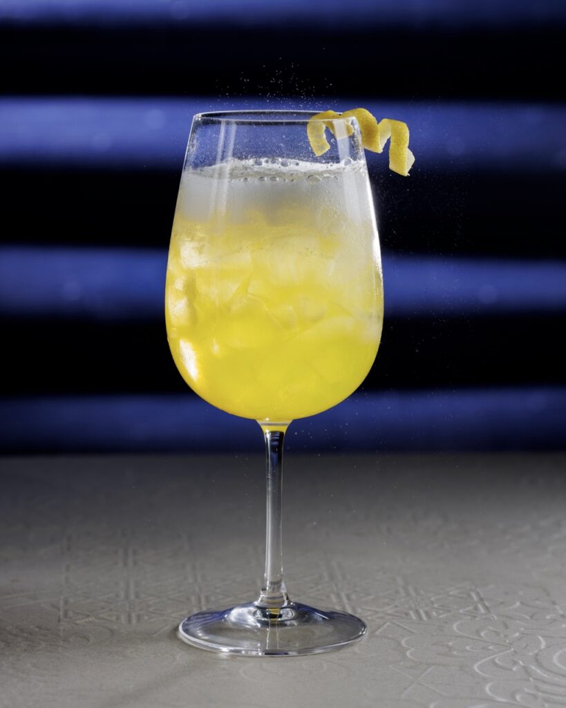 A tall glass with a yellow cocktail with a lemon twist garnish.