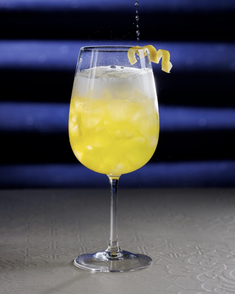 Soda water pours into and bubbles a yellow Emily cocktail in a tall glass with ice and a lemon twist garnish.