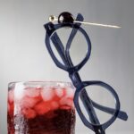 A pair of round glasses lean against a Nigel cocktail in red color with ice cubes as a luxardo cherry on a toothpick balances on the glasses.
