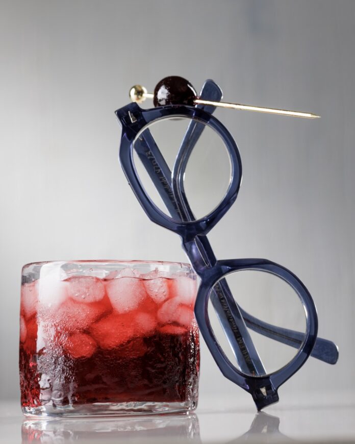 A pair of round glasses lean against a Nigel cocktail in red color with ice cubes as a luxardo cherry on a toothpick balances on the glasses.