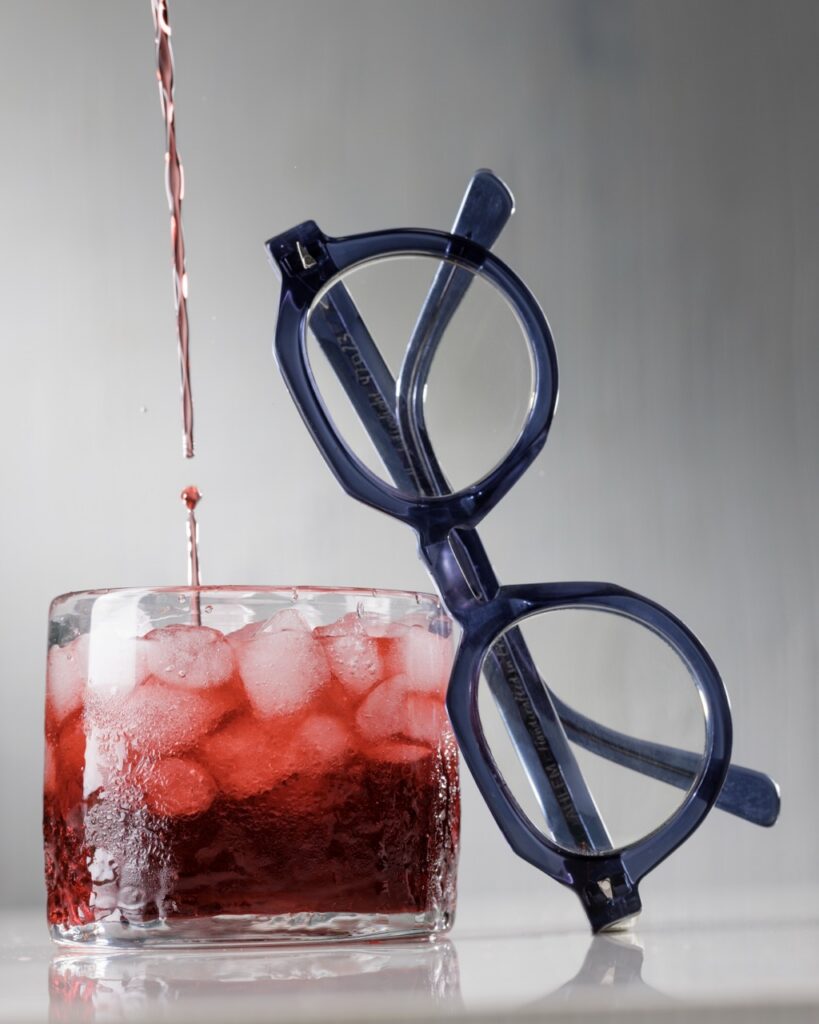 Red liquid pours into a glass with ice that a pair of blue round glasses leans against.